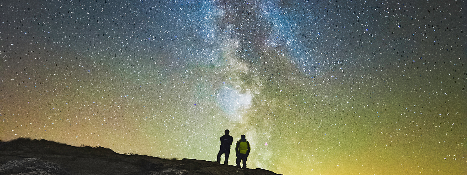 Two people standing on a cliff looking up at the sky full of stars. They're on the west coast of the Island, at a stargazing site in Niarbyl